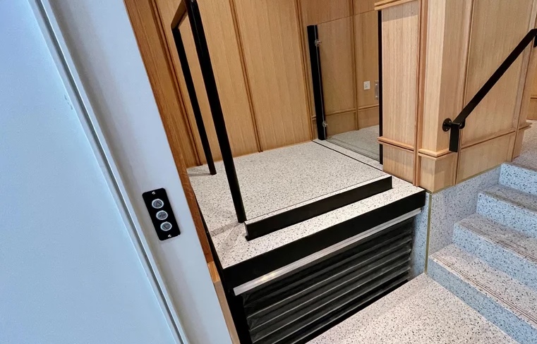 Compact wheelchair lift with black frame, light-colored floor, and wood paneling.