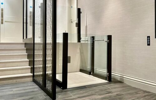 Modern wheelchair lift with glass panels and dark frames next to a staircase.